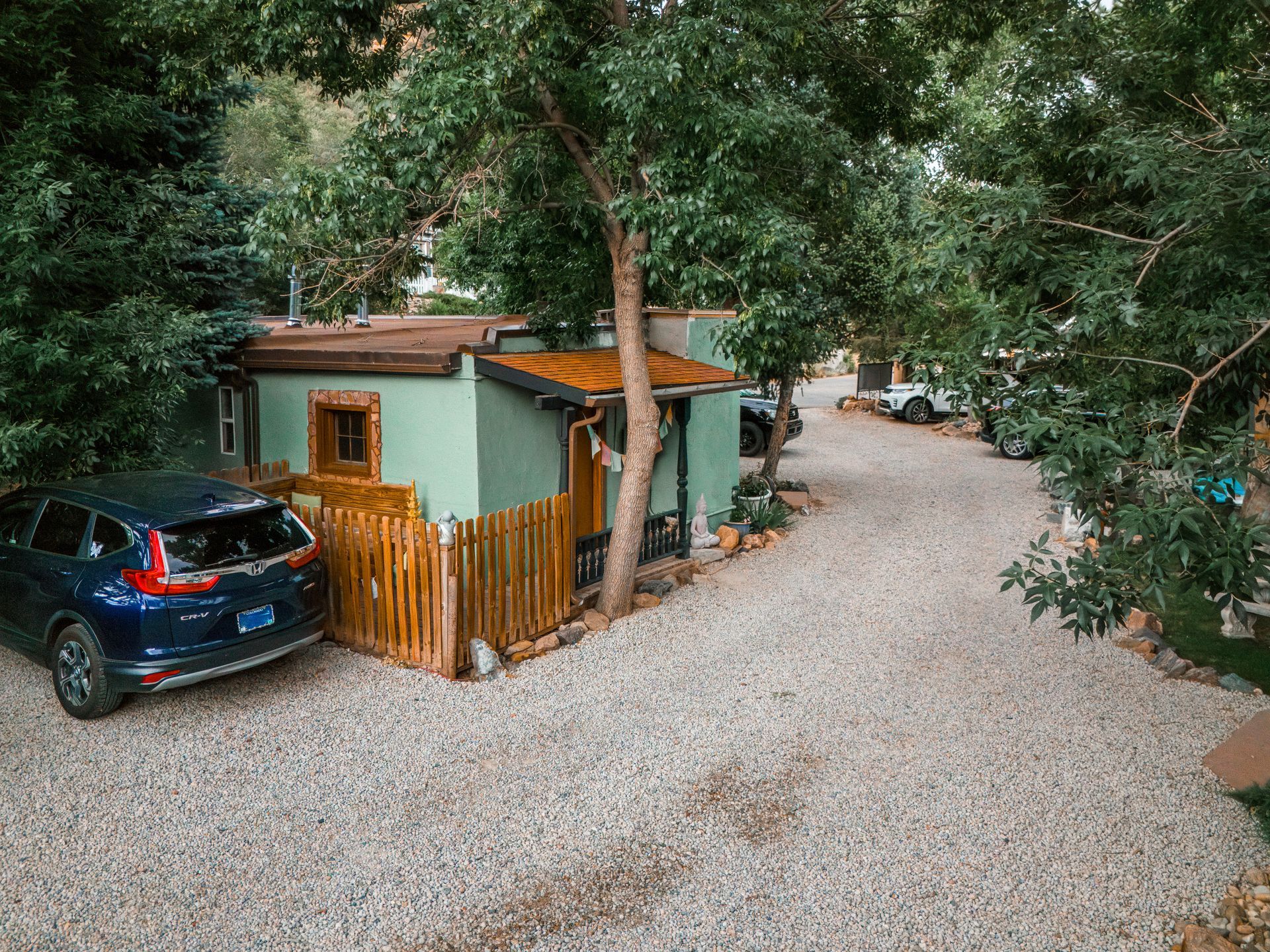 A small, light green cottage with a wood fence and a dark blue SUV parked on a gravel driveway, surrounded by trees.