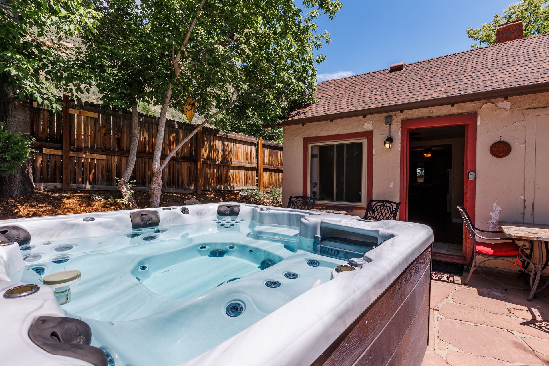 A hot tub filled with clear water sits on a stone patio outside a house next to a wooden fence and leafy trees.