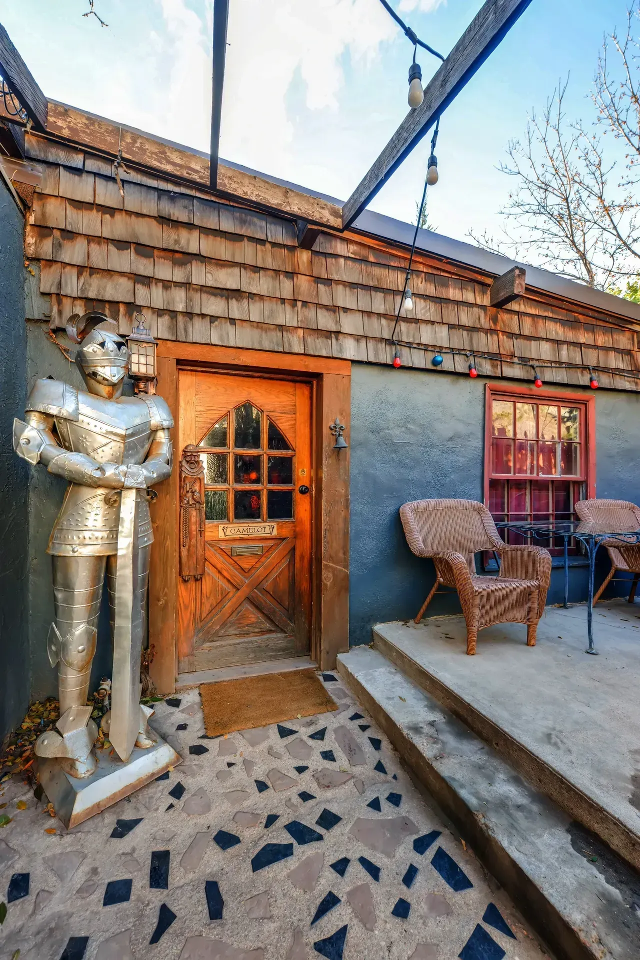 A metal suit of armor stands guard beside a wooden door on a stone patio with wicker chairs under a string of lights.