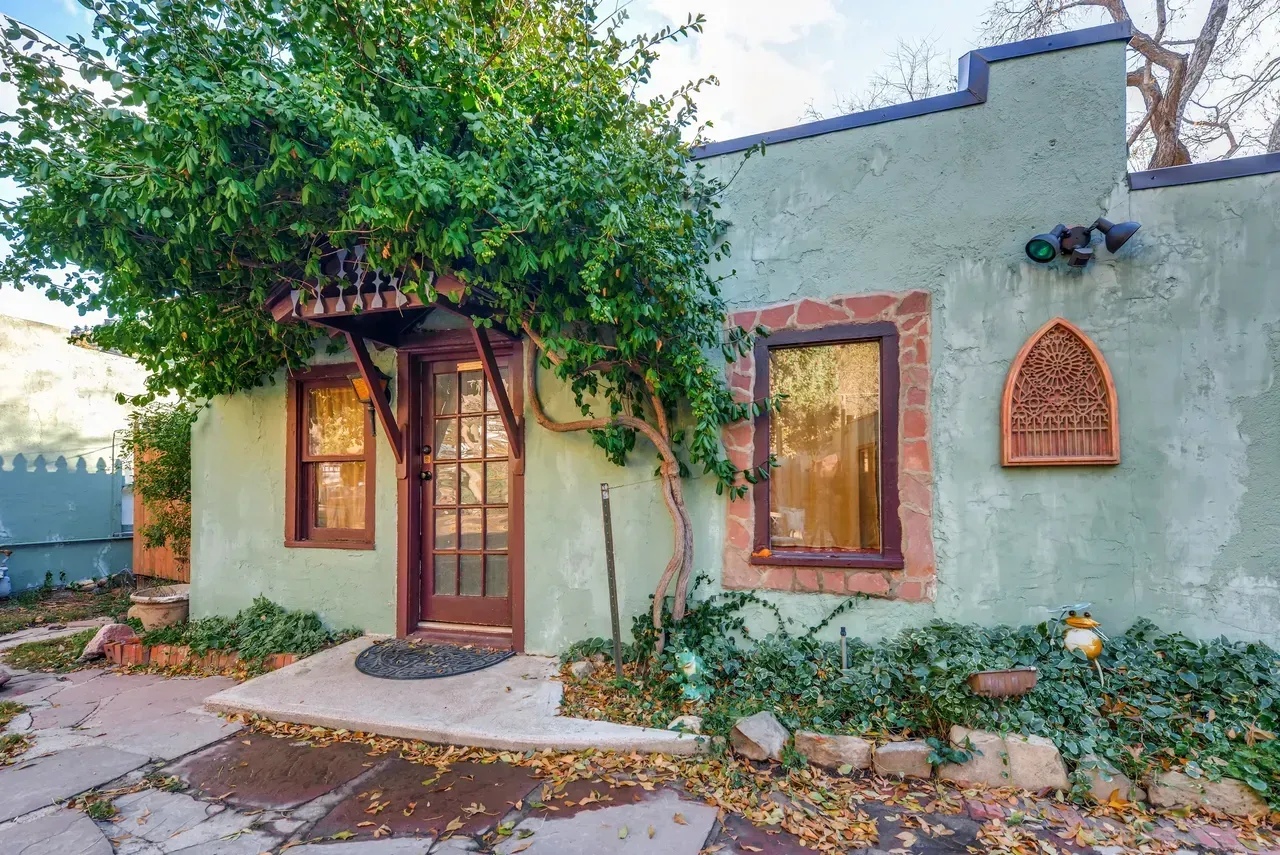 Light green stucco house with a dark wood door, arched window, and a leafy tree canopy, surrounded by garden beds.