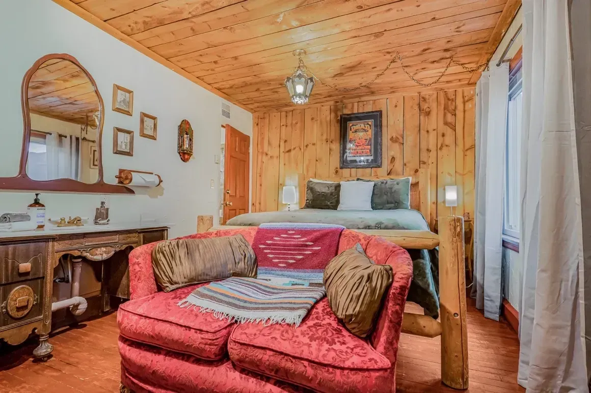 A rustic room features a red sofa in the foreground, a bed with wood-paneled walls and ceiling, and a wooden vanity mirror.