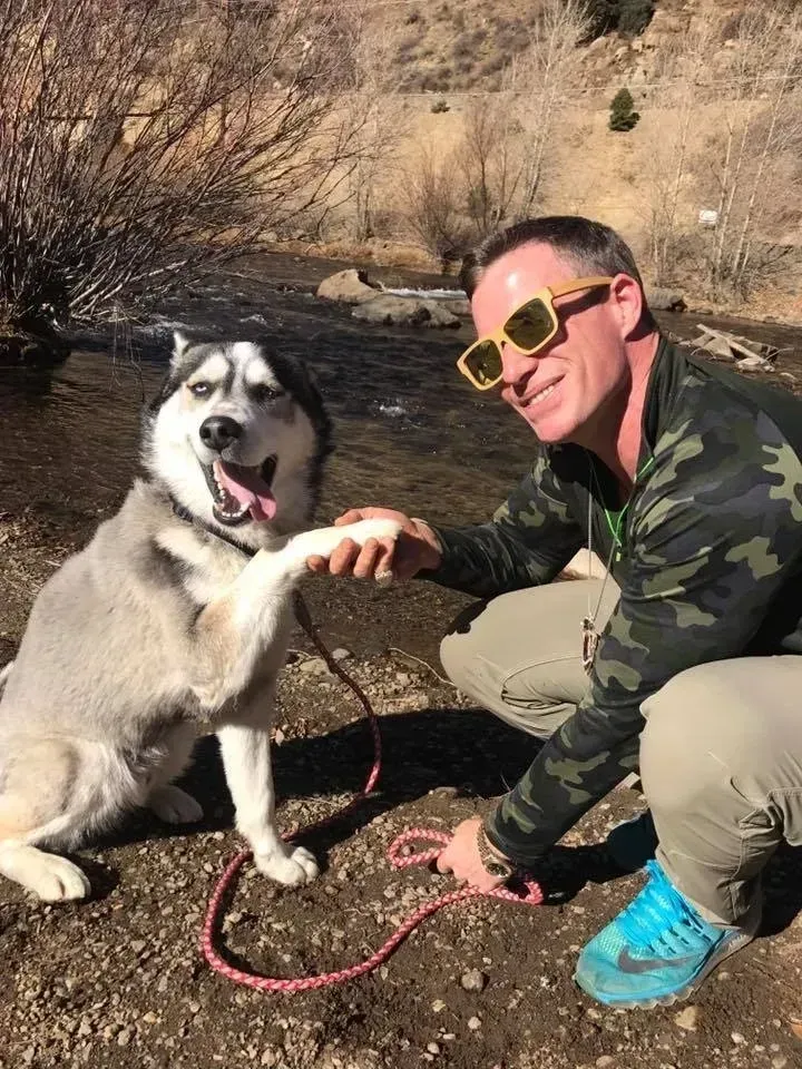 A man in a camouflage top and bright sunglasses holds a husky's paw while crouching by a river.