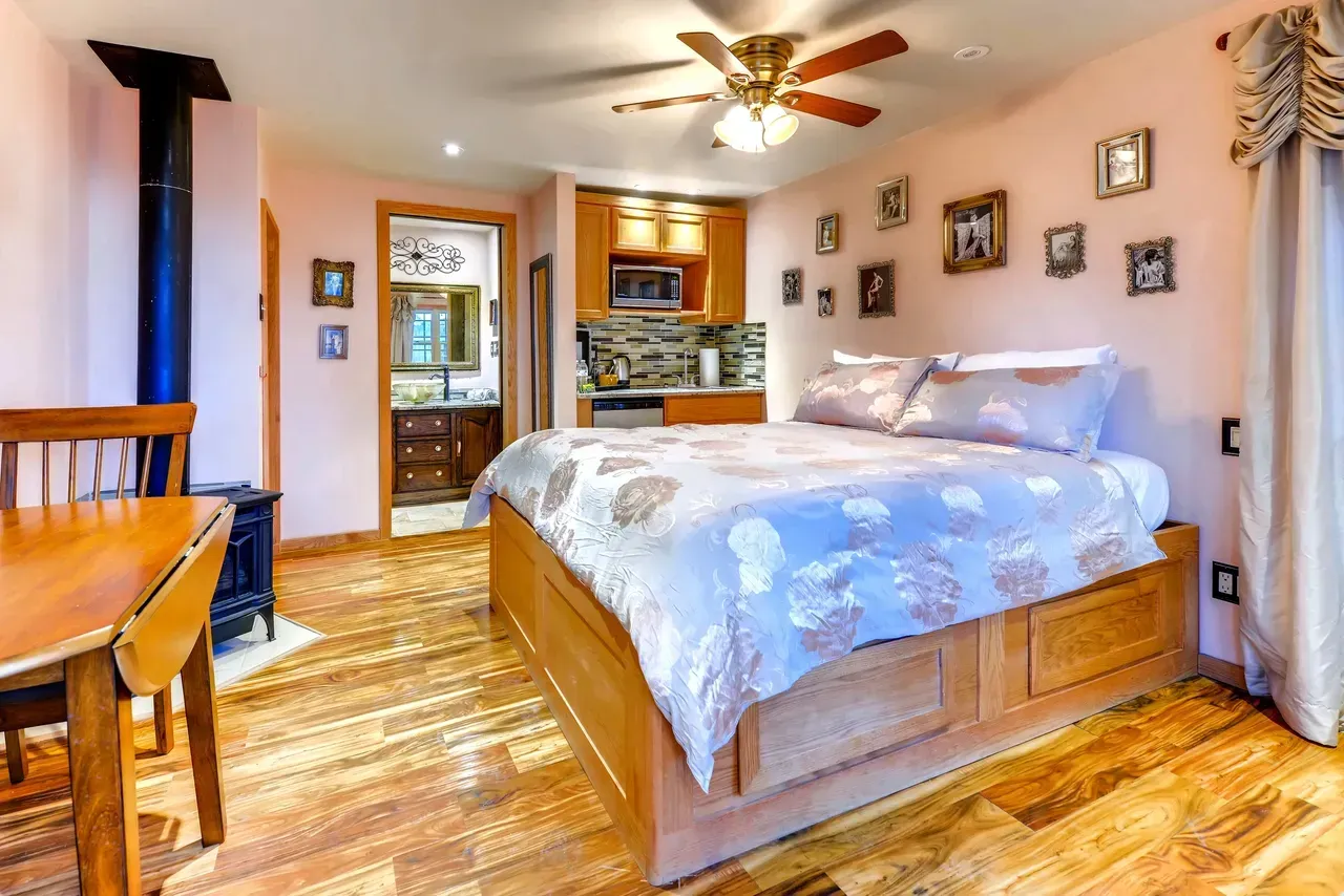 Bedroom with a wooden bed, patterned bedding, light pink walls with framed photos, a small kitchen, and a dining table.