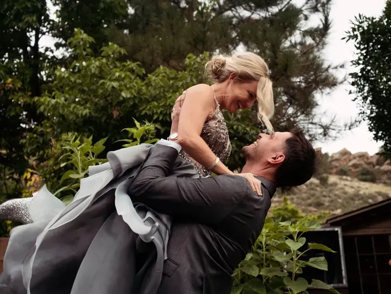 A person in a dark suit lifts and holds a person in a ruffled grey dress outdoors, both smiling at each other.
