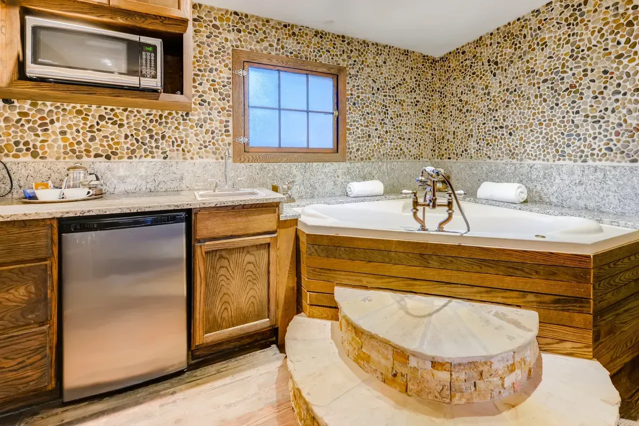 A rustic hotel room with a stone-tiled wall, a small refrigerator, a microwave, and a large jetted bathtub on a raised base.