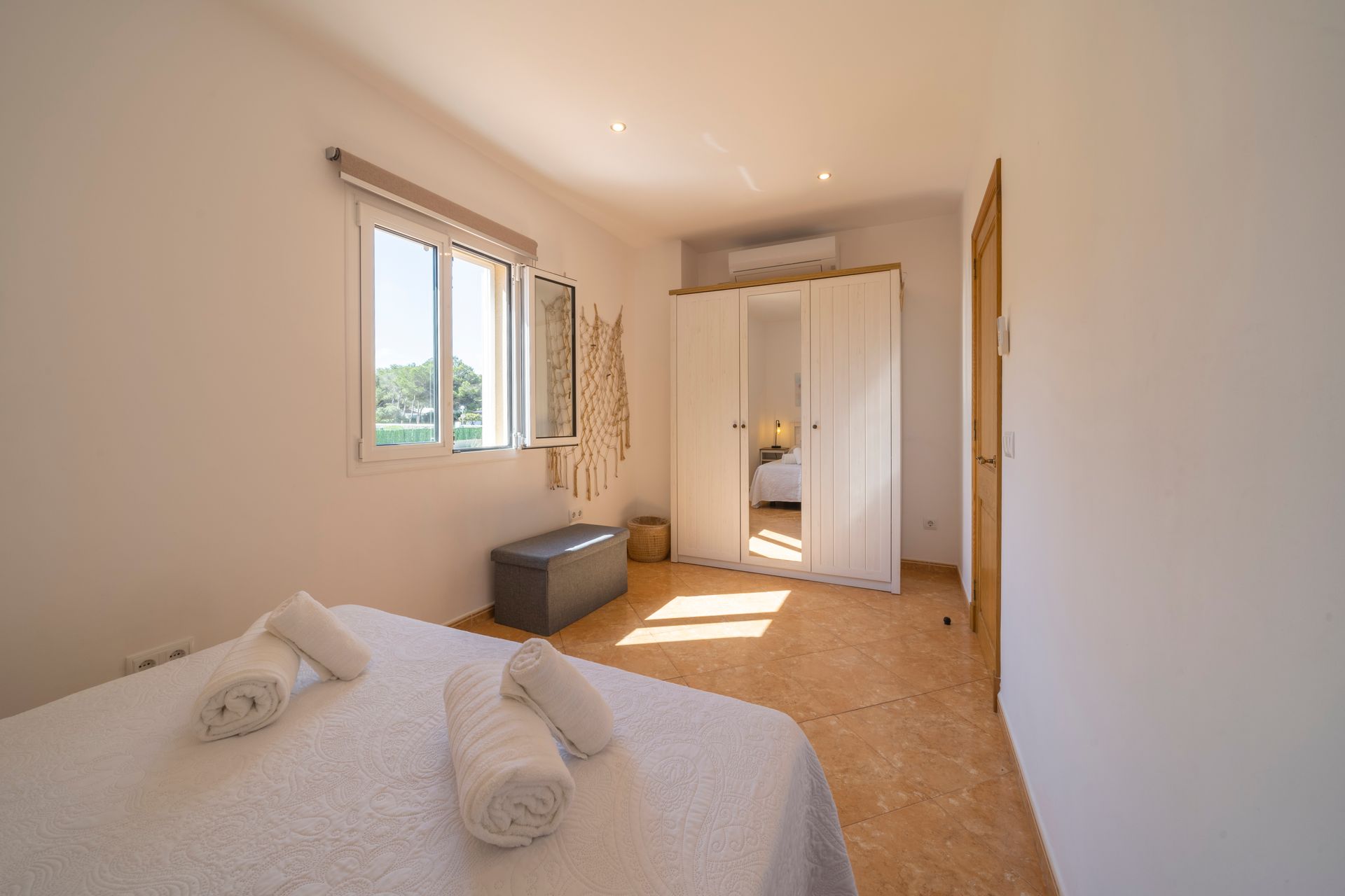 Dormitorio con cama, ventana, armario y suelo de baldosas beige. Entra la luz del sol.