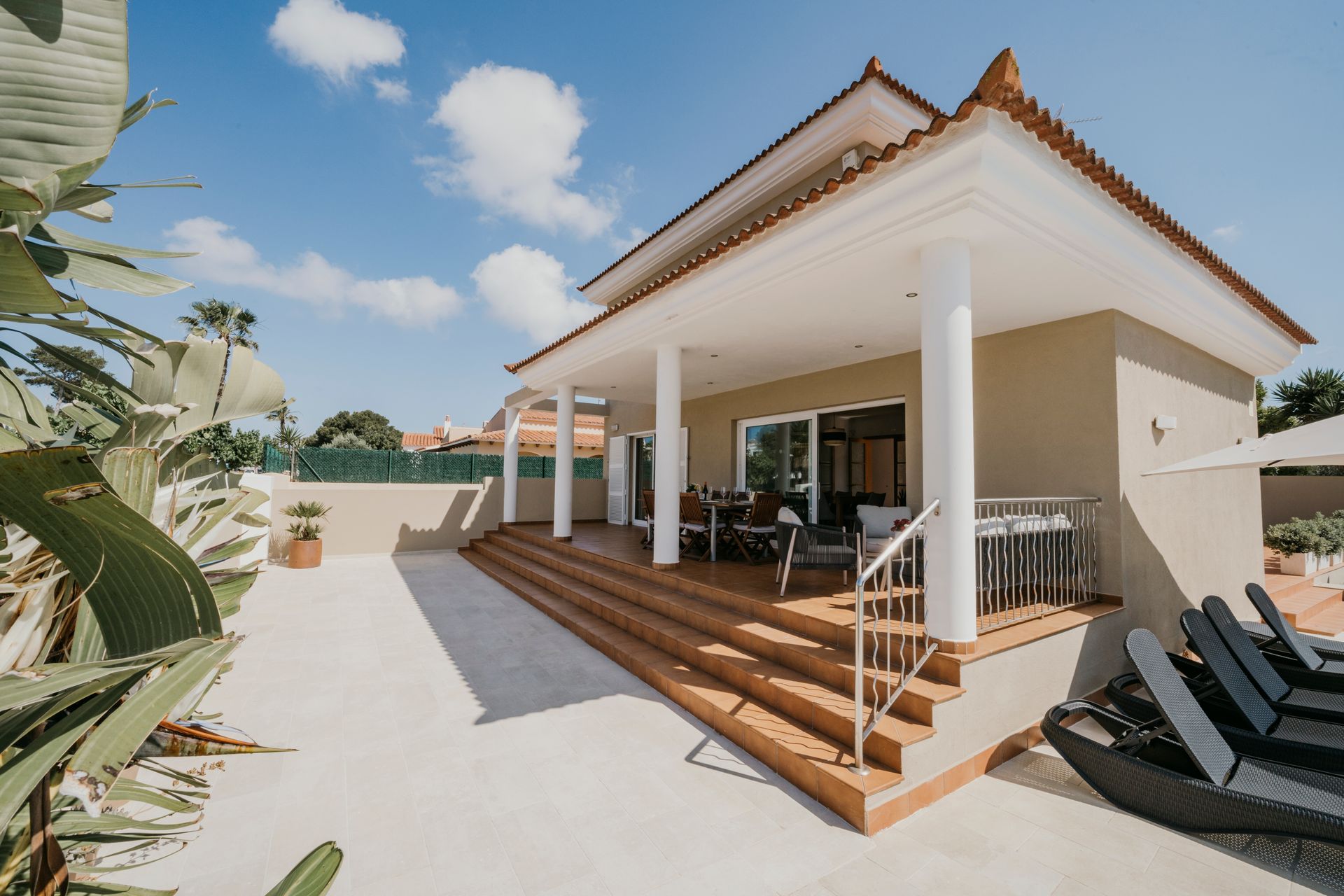 Vista exterior de una villa con pilares blancos, escalones que conducen a un patio y sillones.