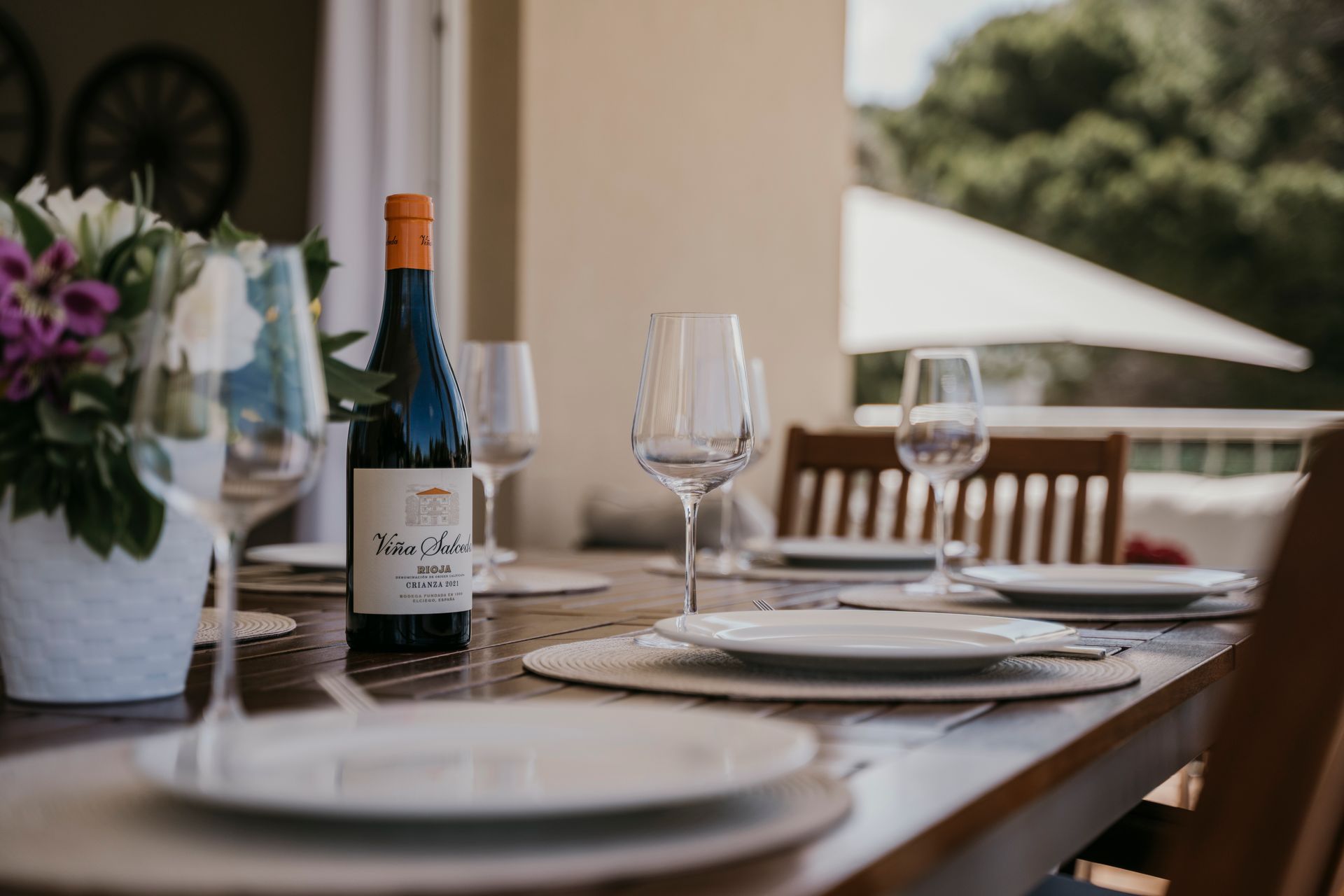 Mesa de comedor al aire libre con vino, copas y platos; flores en jarrón.