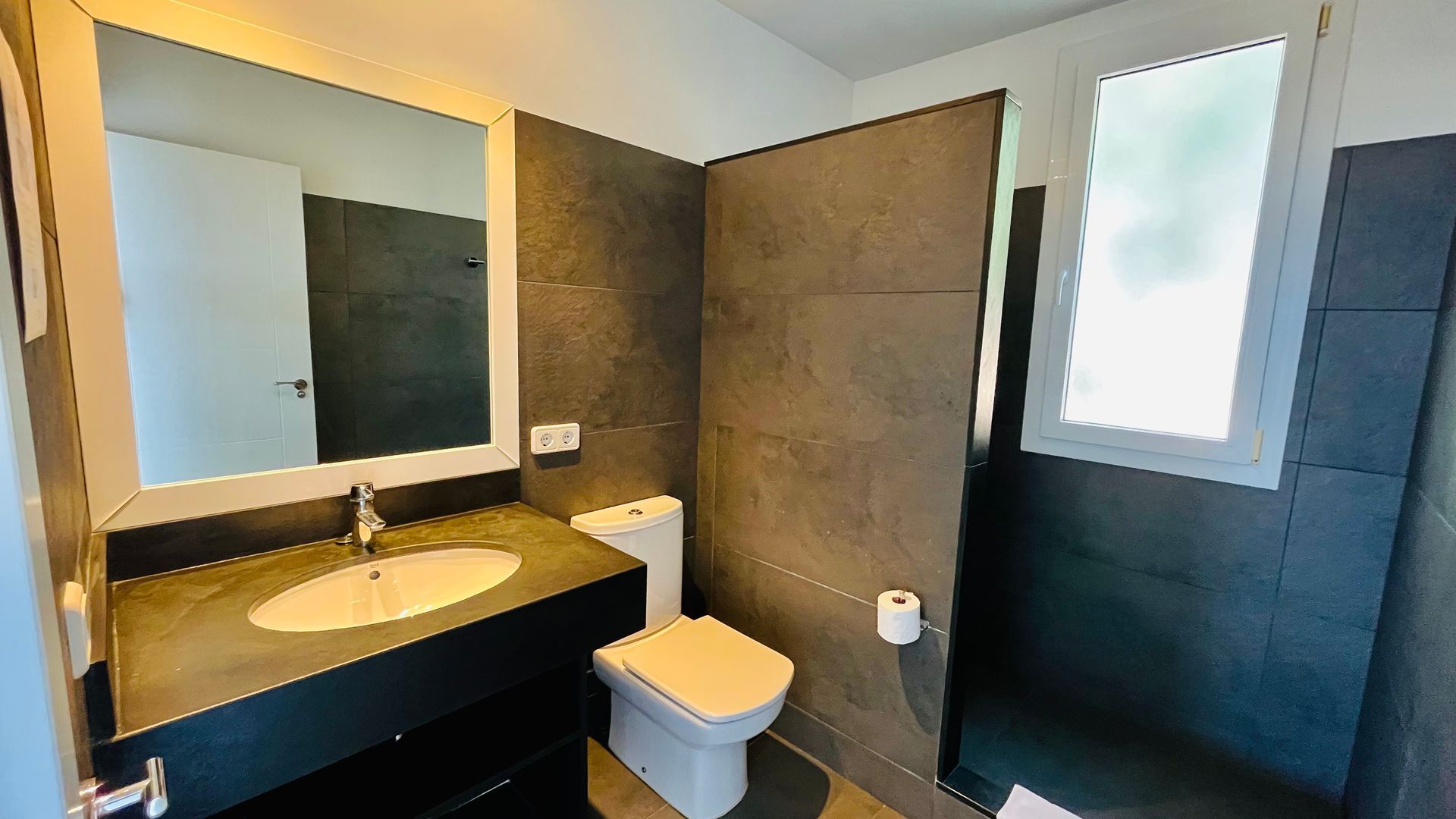Baño moderno con azulejos grises, lavabo, inodoro y ventana.