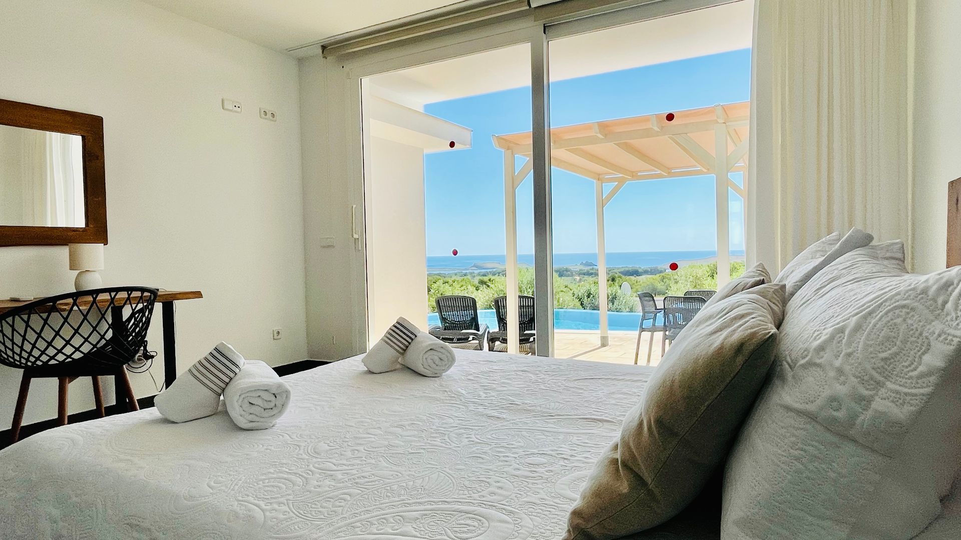 Dormitorio con cama, toallas, escritorio y puerta corrediza de vidrio a patio exterior con piscina y vista.