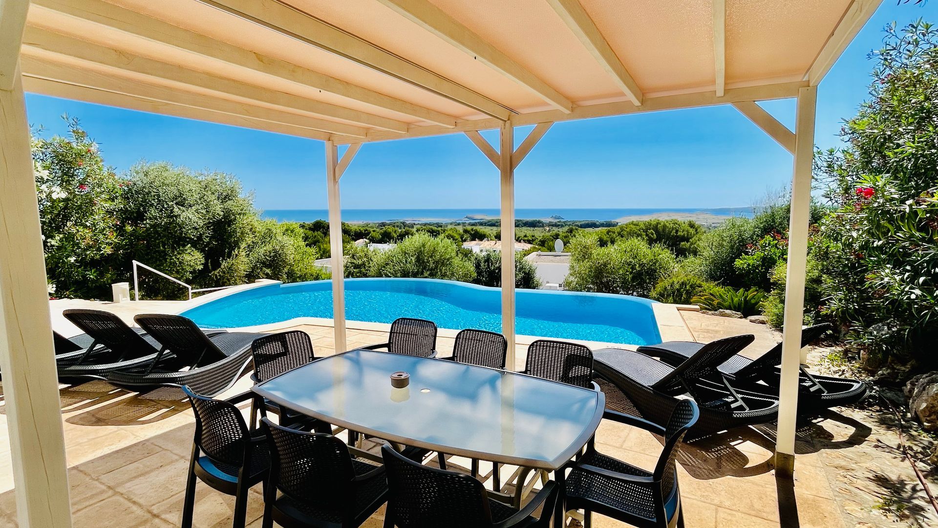 Mesa de comedor al aire libre bajo una pérgola con vista a una piscina, con el océano al fondo.