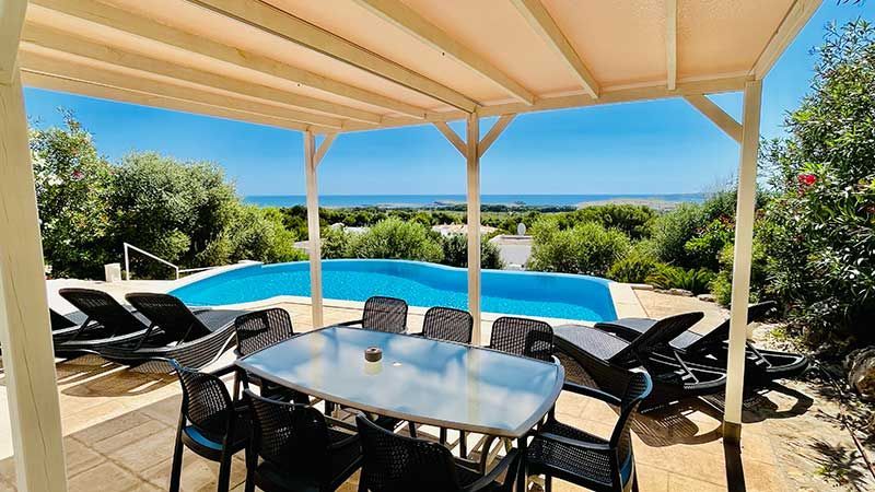 Mesa de comedor al aire libre bajo una pérgola con vista a una piscina, con el océano al fondo.