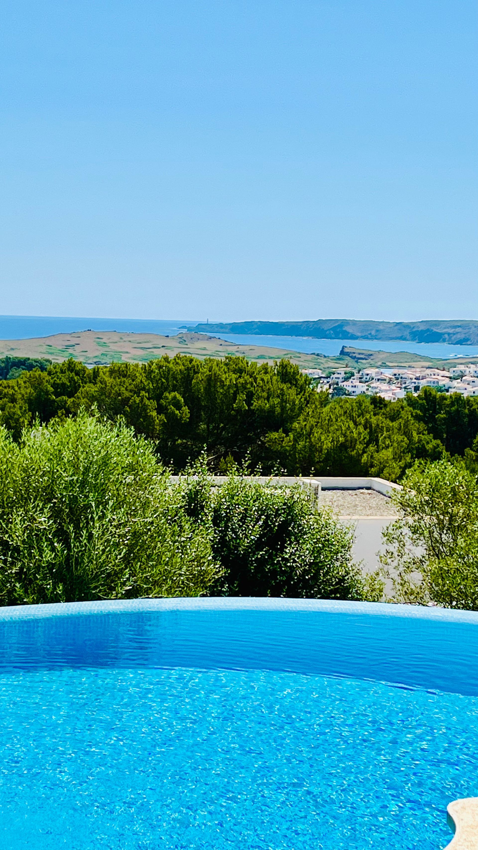 Piscina infinita con vistas a un pueblo costero y al mar bajo un cielo azul claro.