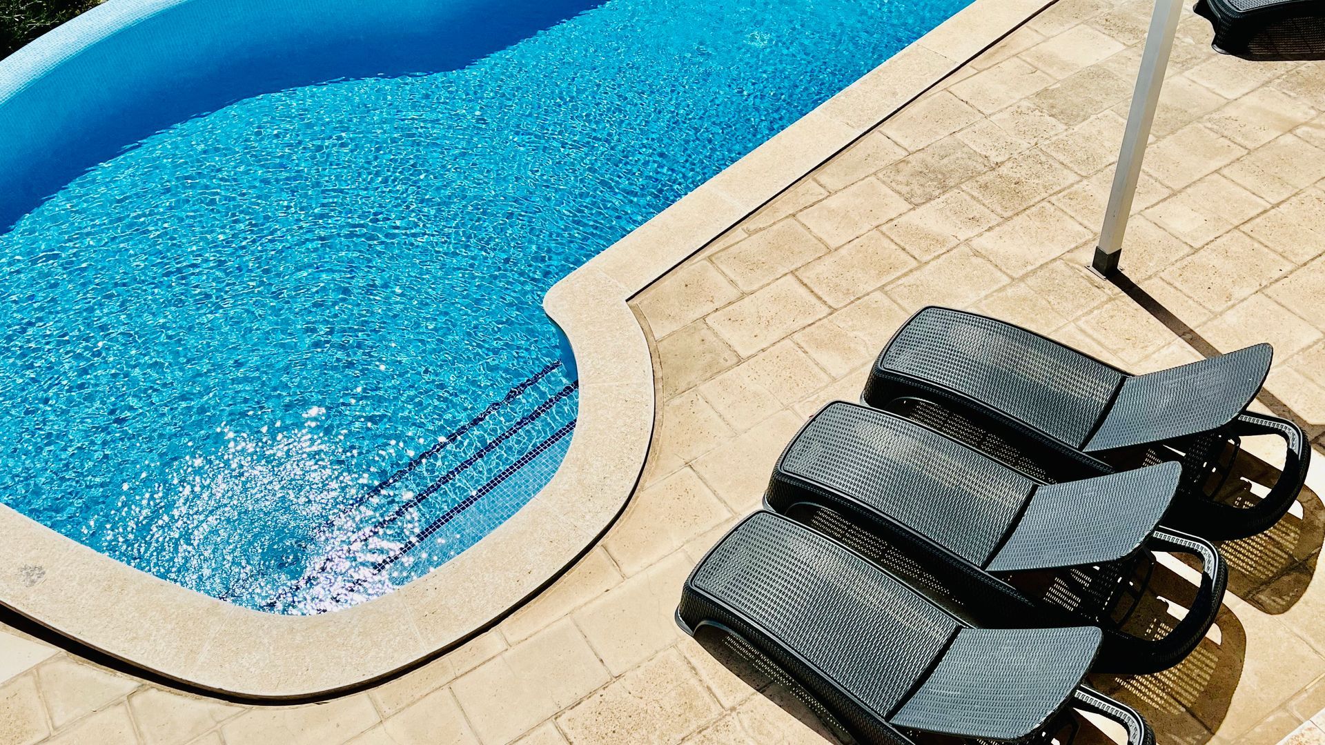 Escena junto a la piscina: piscina azul, patio beige, tres tumbonas grises.
