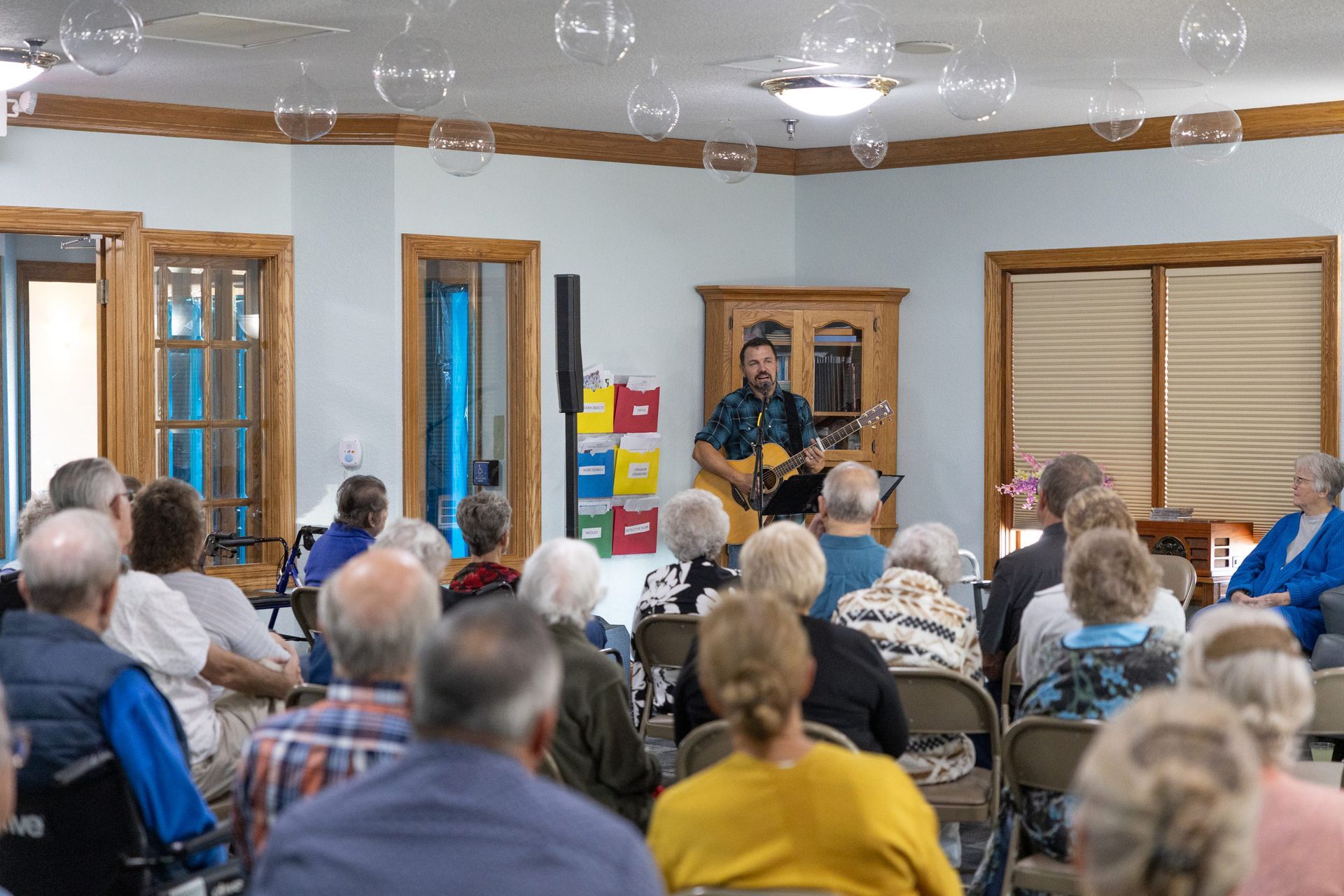 Man playing guitar for a seated audience in a room with windows, light blue walls, and decorations.