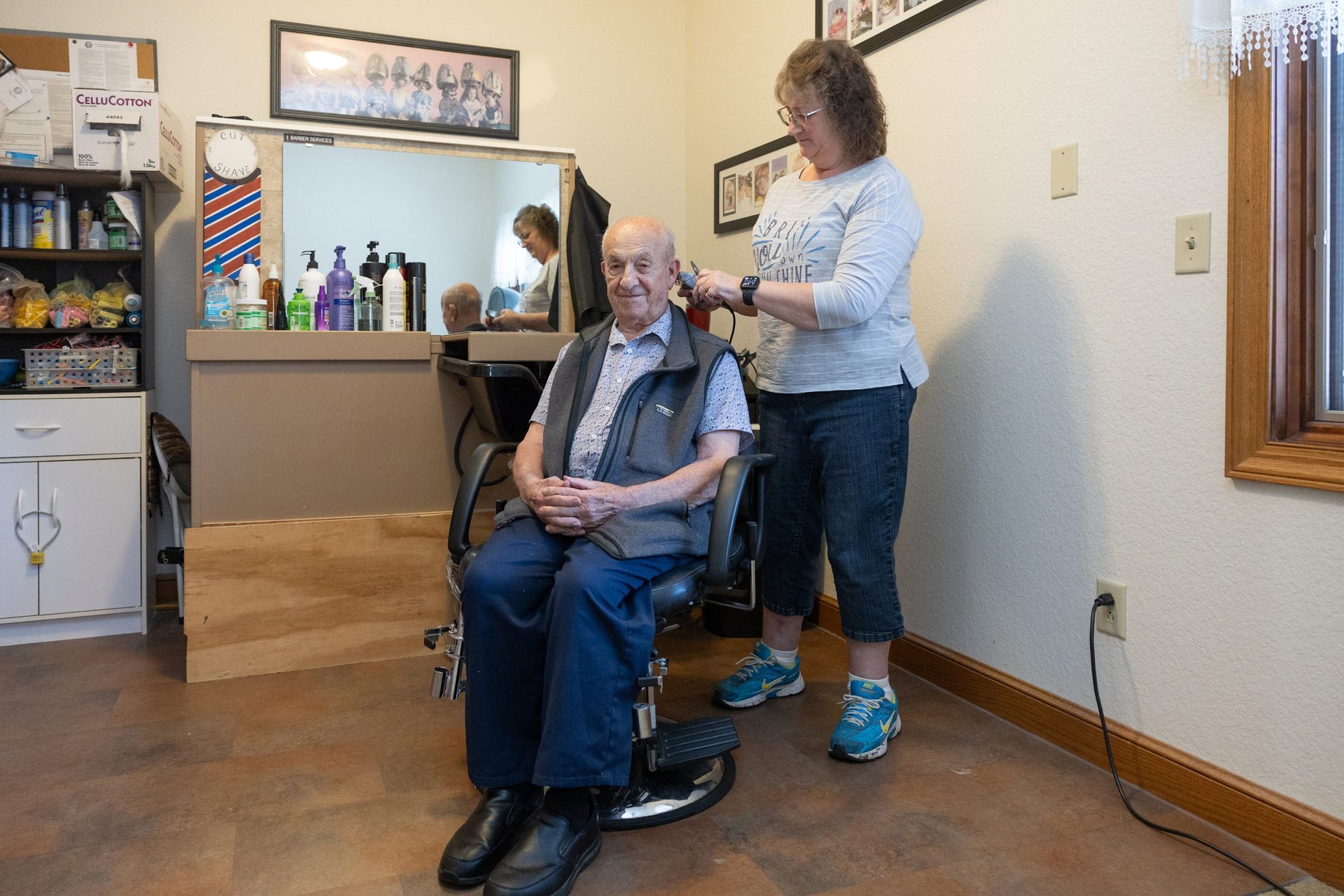 Woman giving a haircut to an elderly man in a wheelchair. They're indoors, near a mirror.