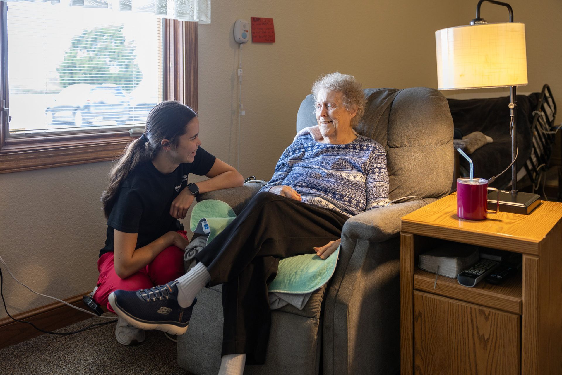 Young woman tending to elderly woman seated in recliner, indoors.