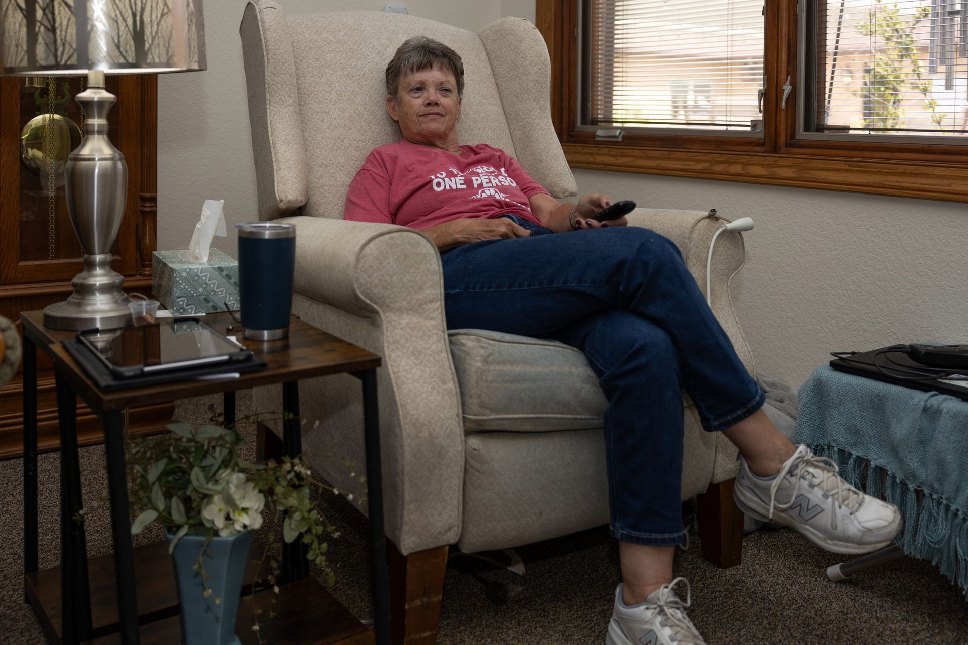 Woman sits in a beige armchair, holding a remote. She wears a pink shirt and blue jeans in a living room.