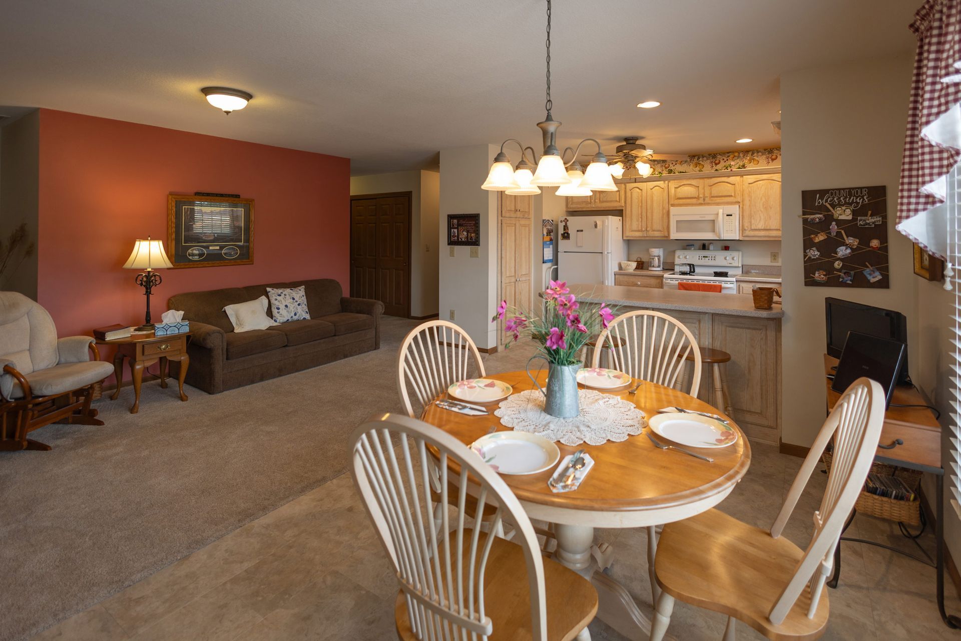 Cozy open-concept living space with dining table, kitchen, and seating area. Red accent wall, beige carpet.