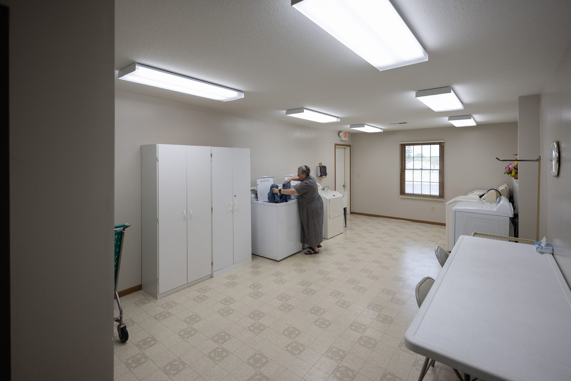A well-lit laundry room with a woman loading a washing machine, white cabinets, and a folding table.