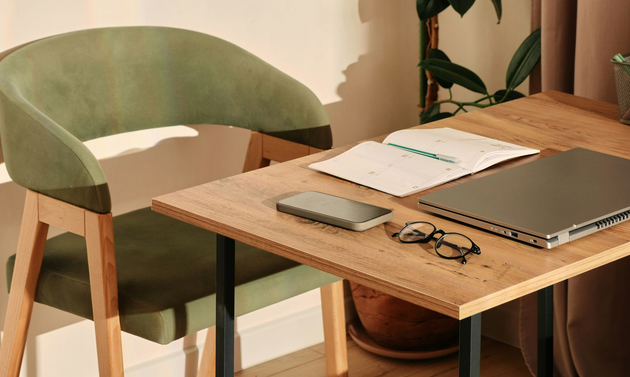 Green chair beside a wooden desk with laptop, notebook, phone, and glasses in a warm-lit room