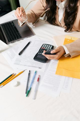 Person calculating on calculator at desk with laptop, papers, pens, and yellow folder