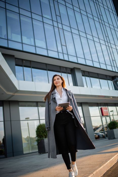Woman walking outside a modern glass office building, holding a phone and wearing a long coat.