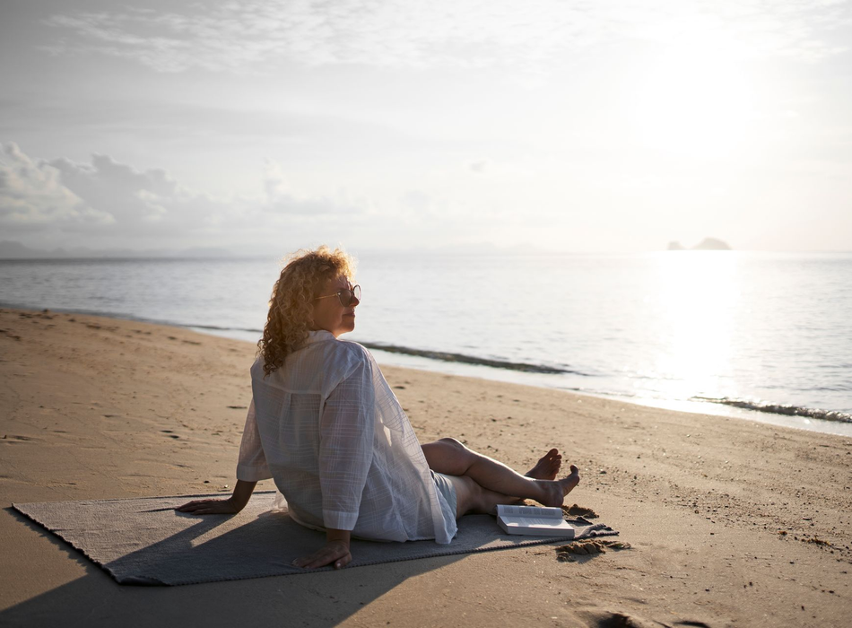 Person sitting on a mat at a sunny beach, facing the ocean at sunrise.