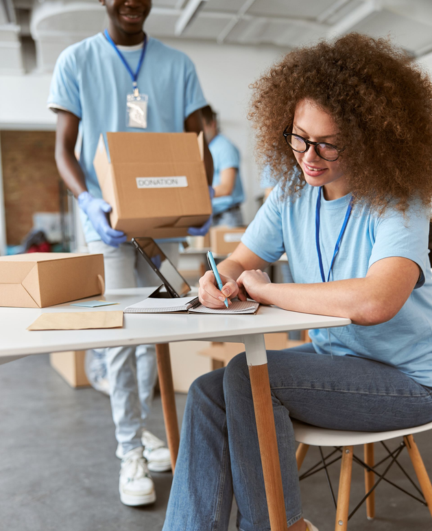 Volunteer labeling boxes at a table while a seated colleague writes in a bright workspace.