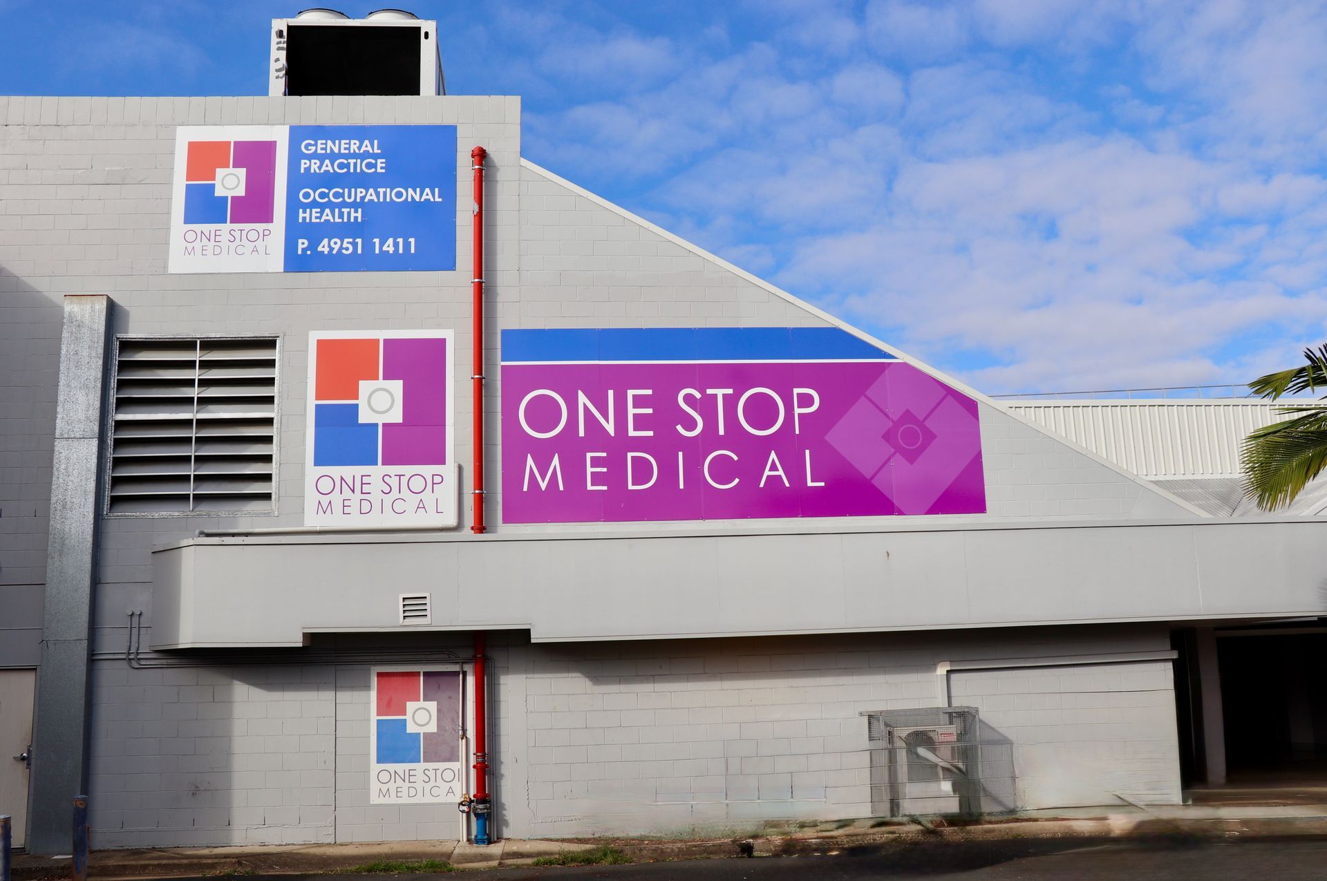 Exterior of One Stop Medical building with several signs. Gray building, blue sky, purple, red, and white logos.— One Stop Medical in Mackay, QLD