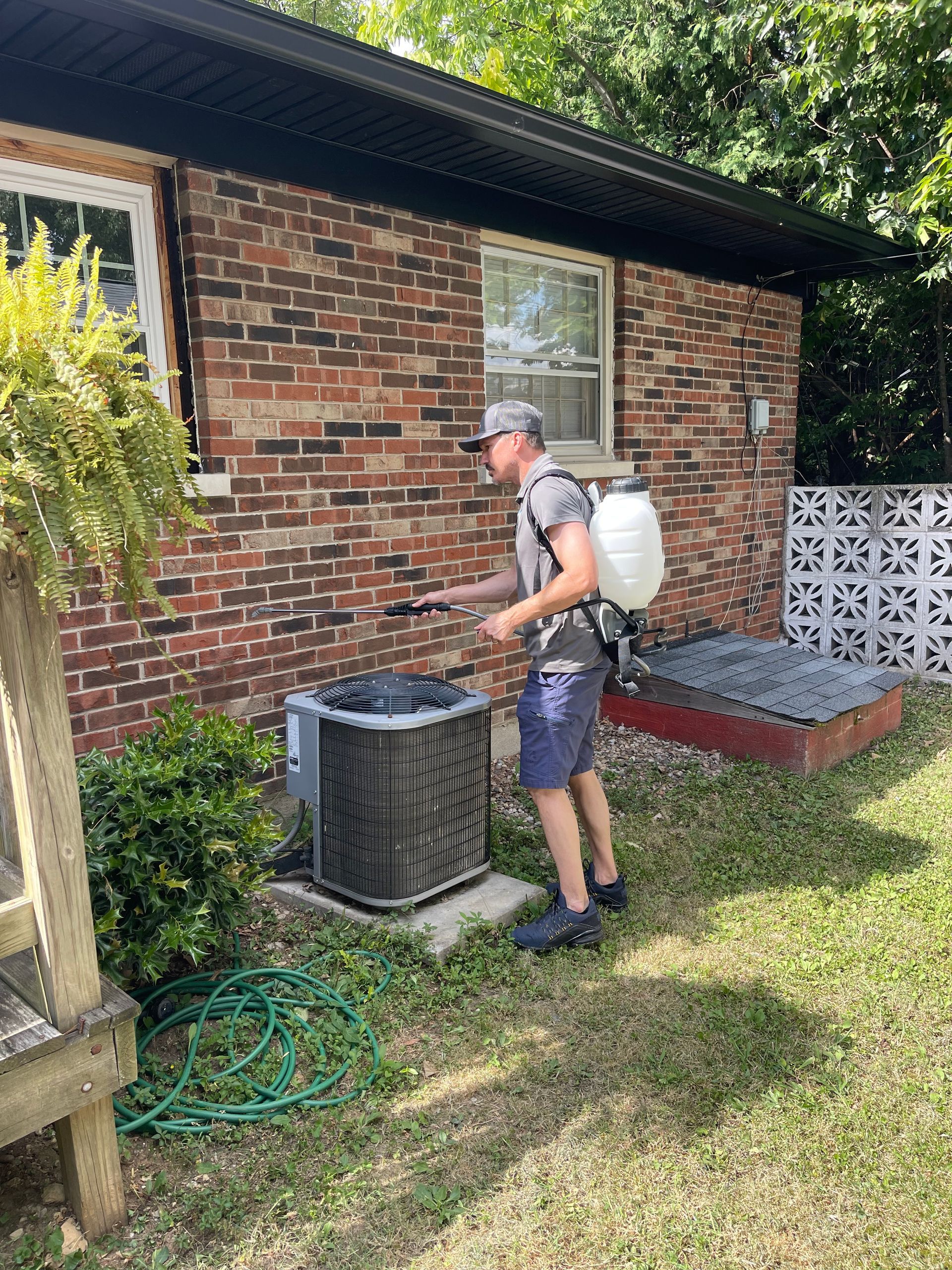 Person spraying an air conditioning unit with a backpack sprayer outside a brick building.