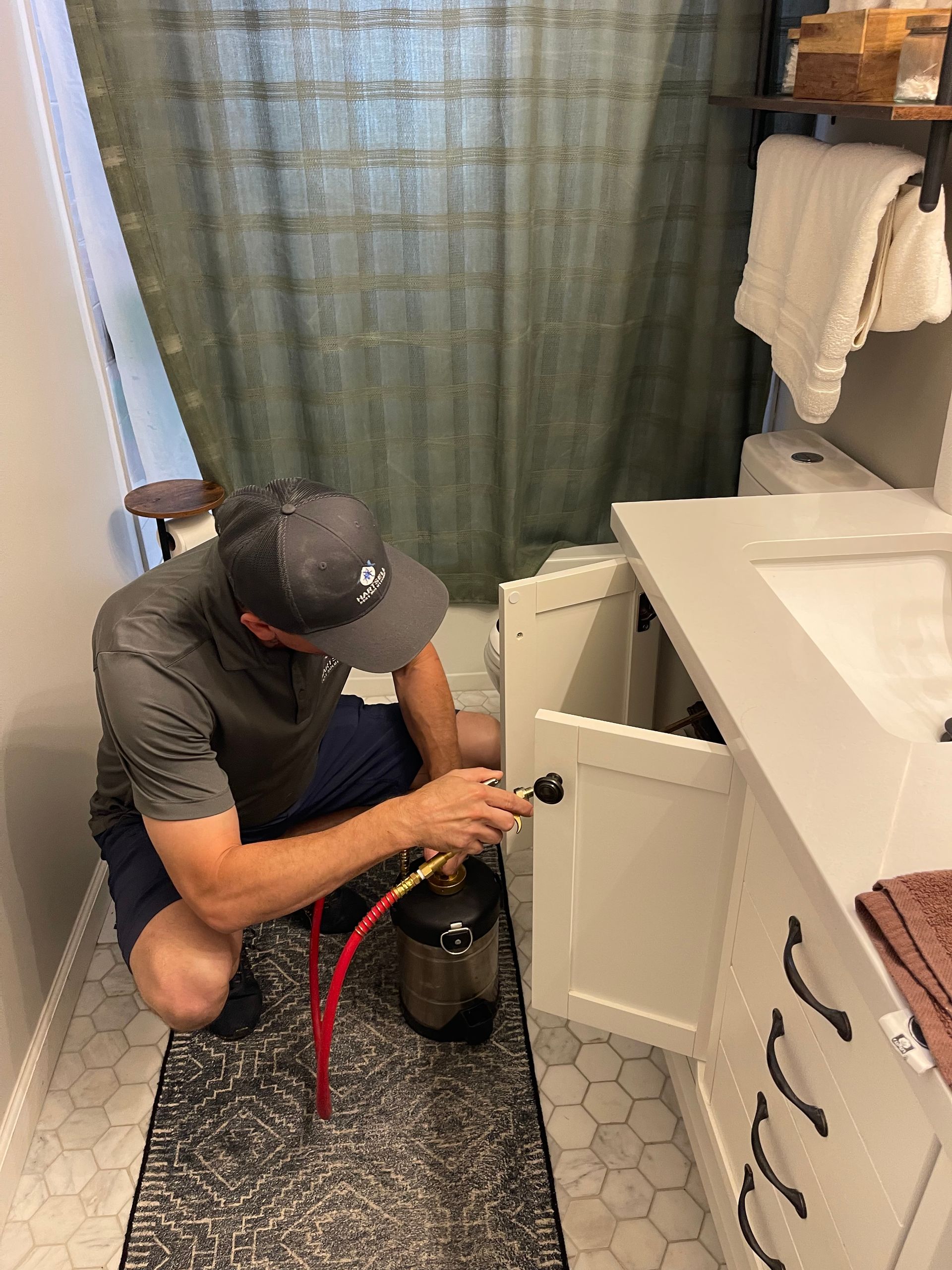 Man kneeling by a white bathroom vanity, using a canister with a red hose.