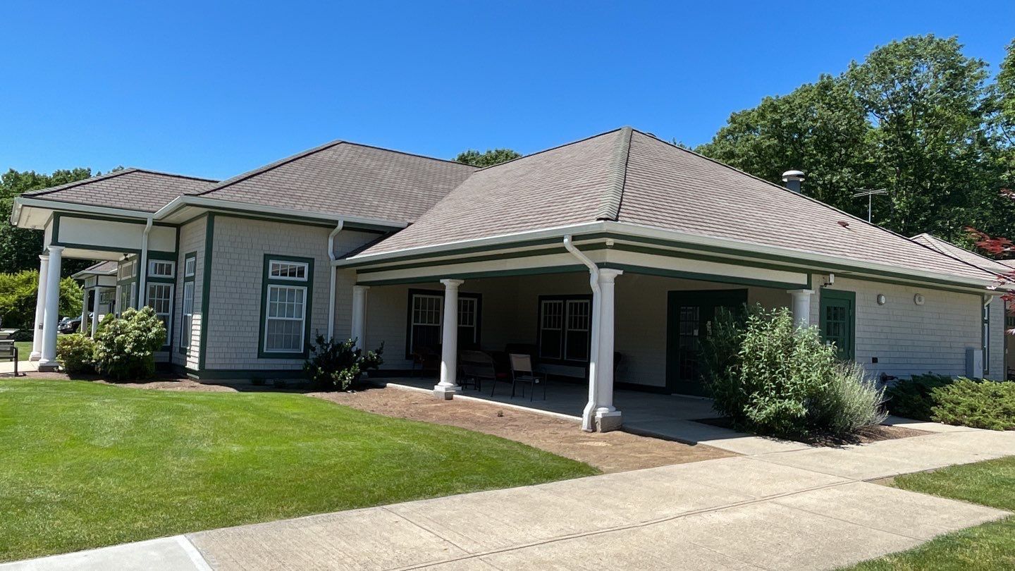 Low-slung building with a wraparound porch, green trim, and a gray roof under a bright blue sky. Green grass and a sidewalk lead up to the porch.