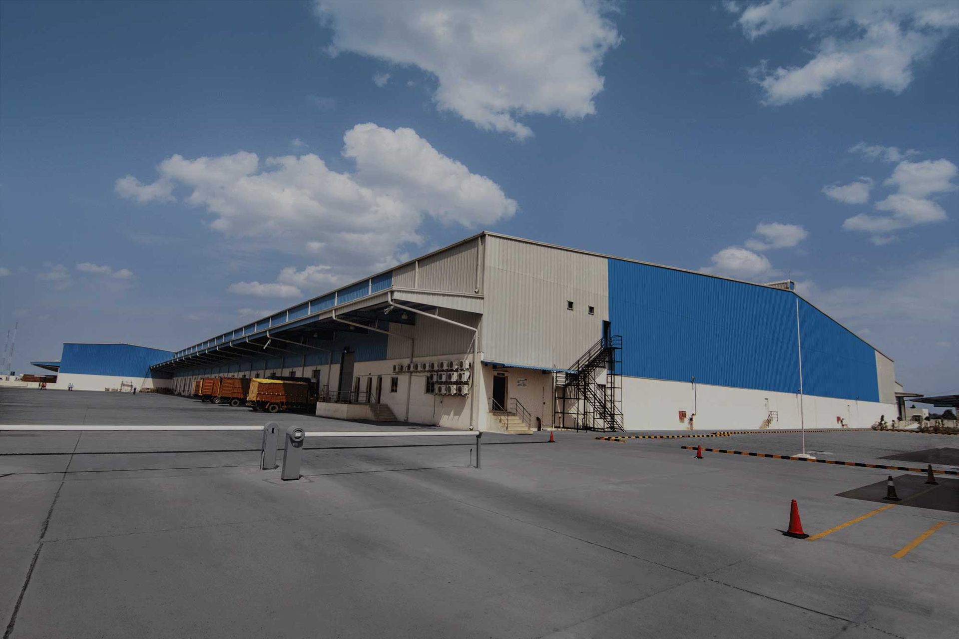 Large warehouse with blue and white panels under a partly cloudy sky. Yellow trucks are parked at loading docks.