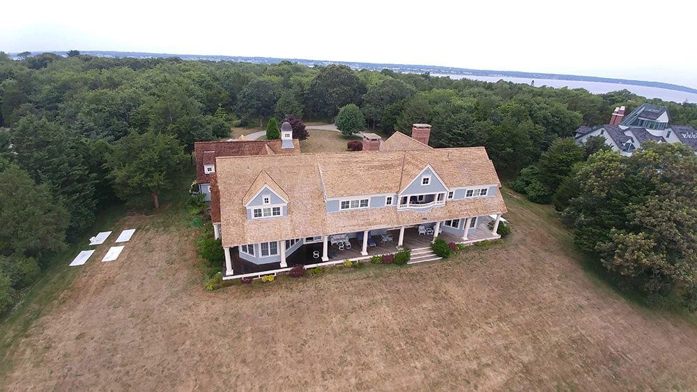 Aerial view of a large, grey house with a brown shake roof and a long porch, surrounded by trees and a dry lawn.