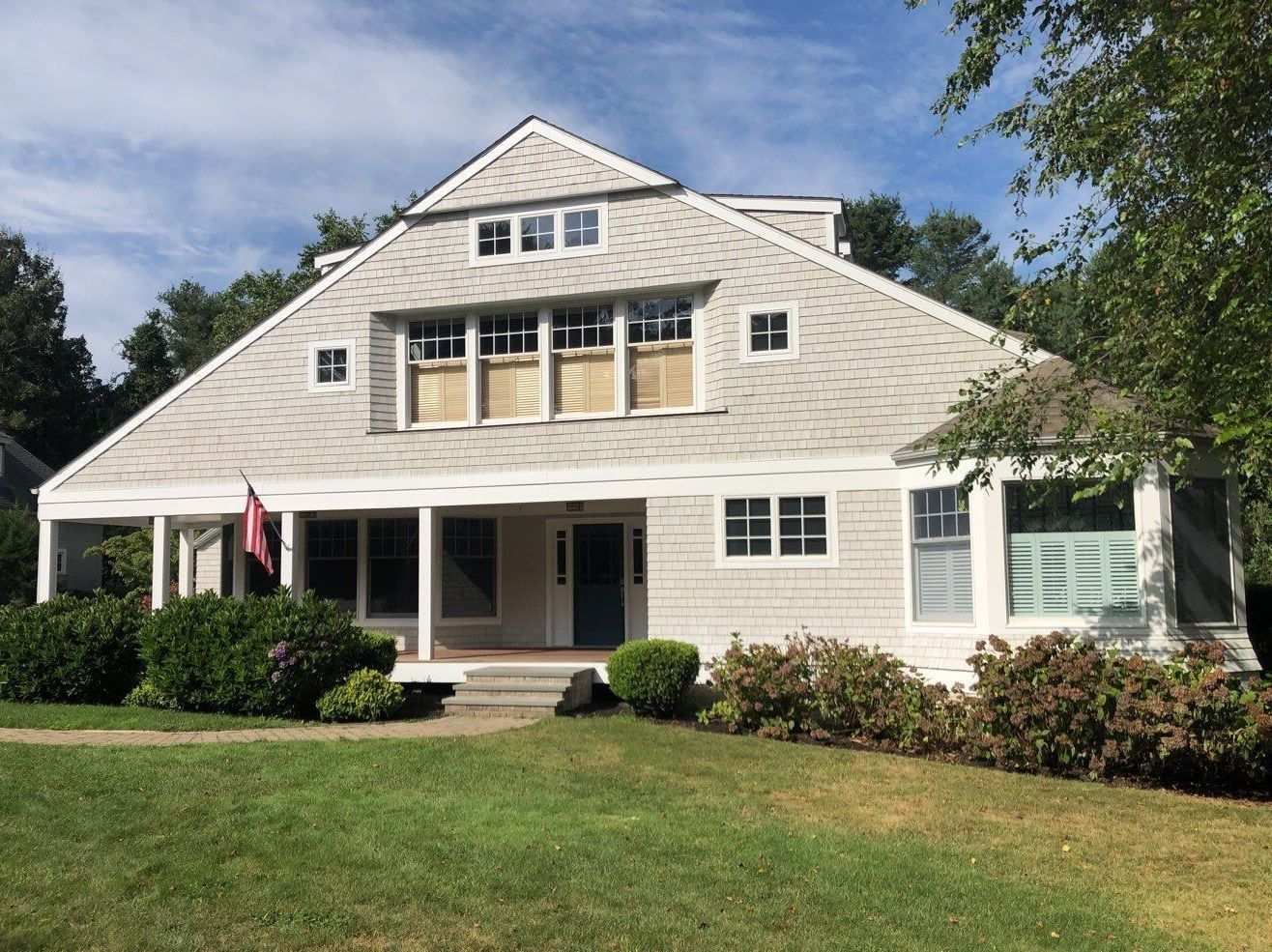 A light gray house with a gabled roof, porch, and windows. A blue door, green lawn, and bushes are in the foreground.