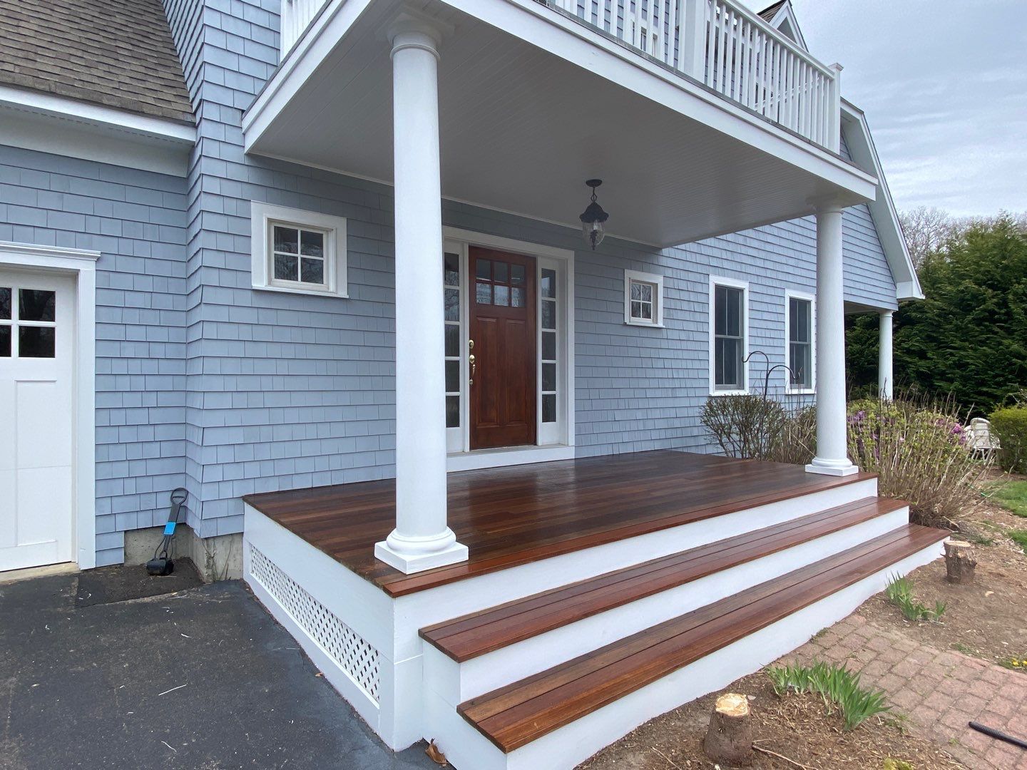 A house with a wooden porch, steps, and blue siding. The porch has white columns and a brown door, with a second-story balcony.