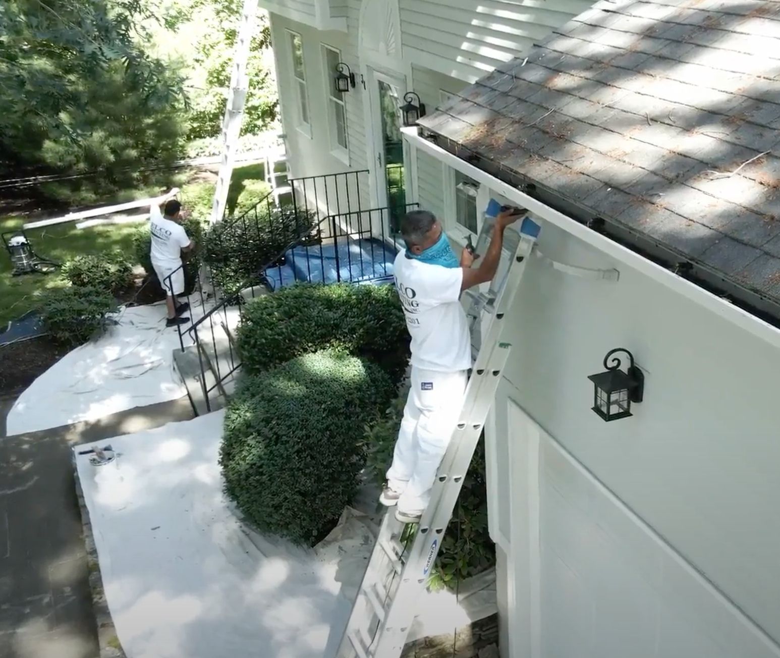 Two painters in white coveralls are working on a house exterior. One is on a ladder near the roofline. The house is white.