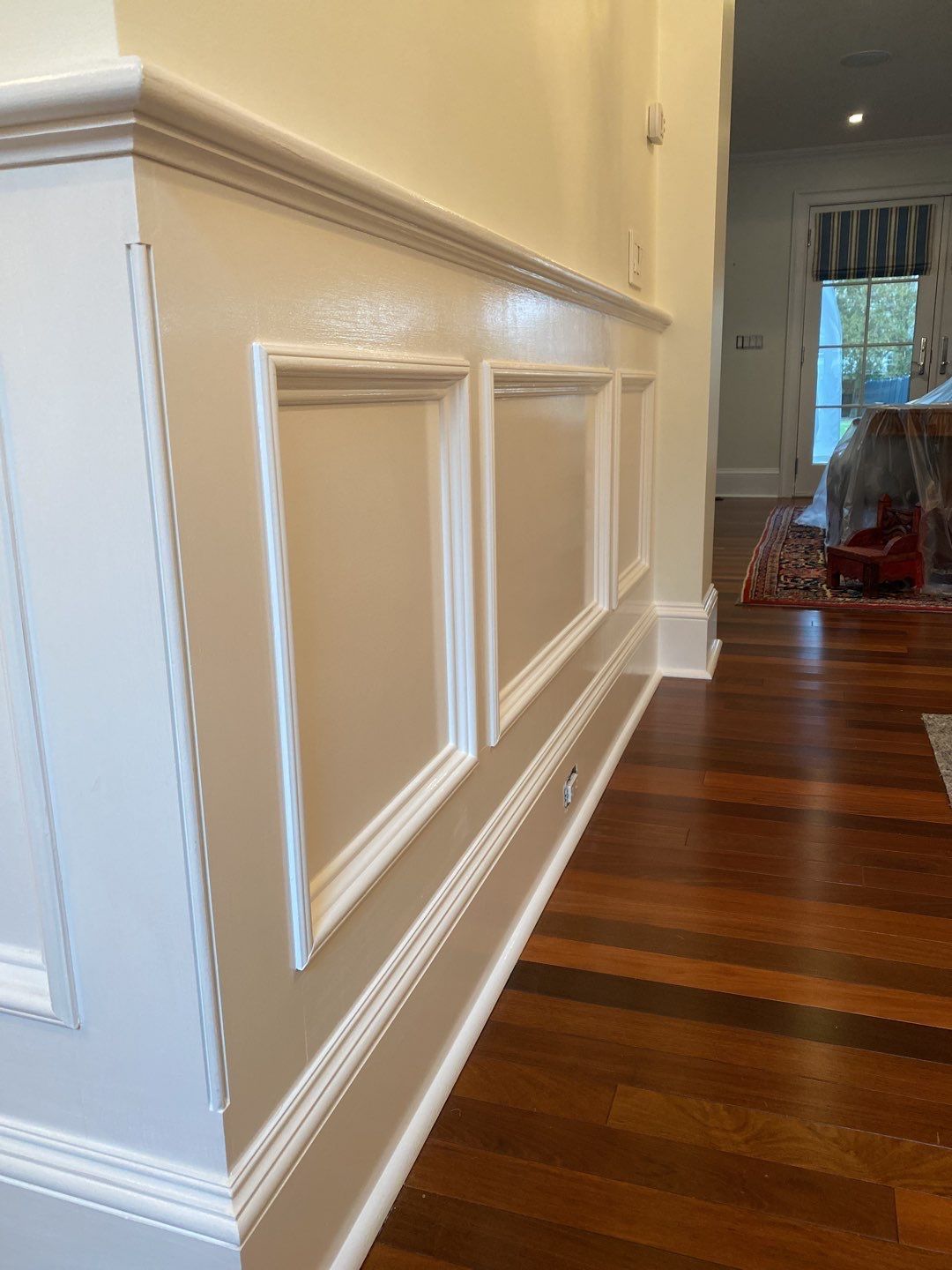 Cream-colored wainscoting with rectangular panels on a beige wall, bordering dark wooden floors. White trim defines the panels and the walls.