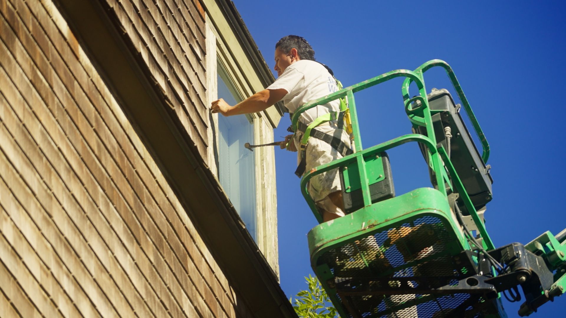 Man in a safety harness on a green lift, working on a window on a brown wooden house exterior. Blue sky.