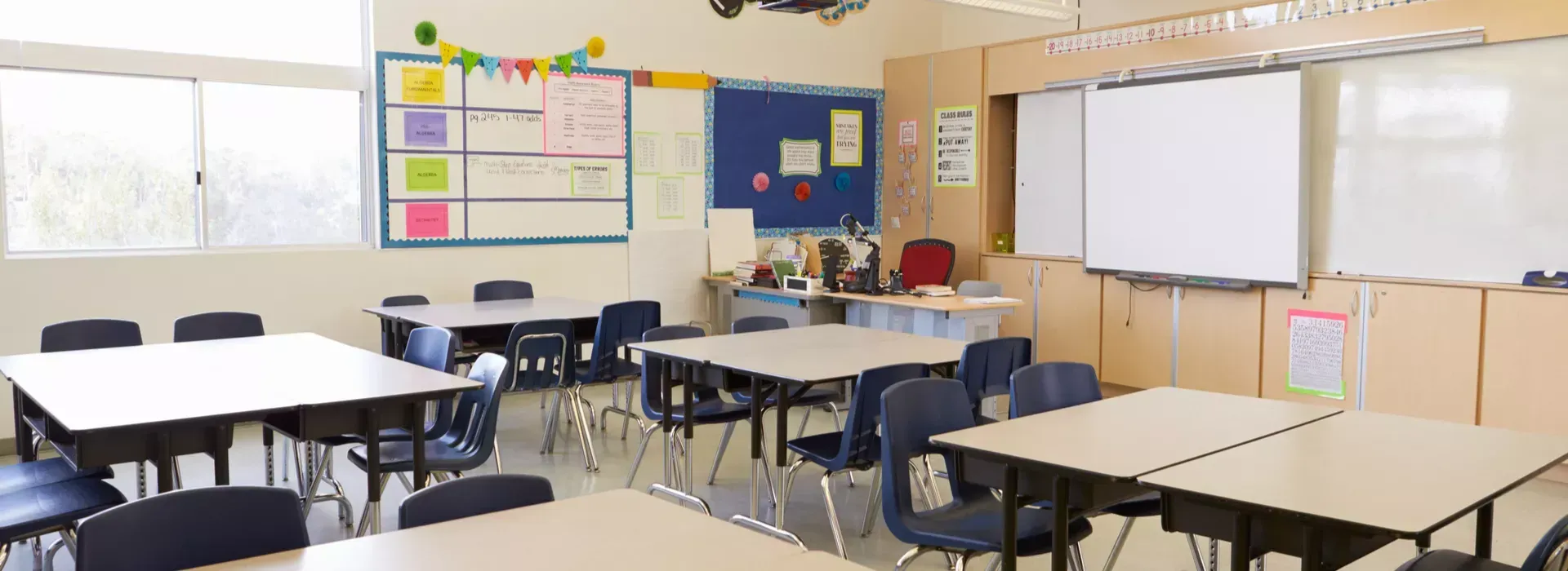 A classroom with tables and chairs, a whiteboard, and colorful decorations. The setting is bright and likely for young students.