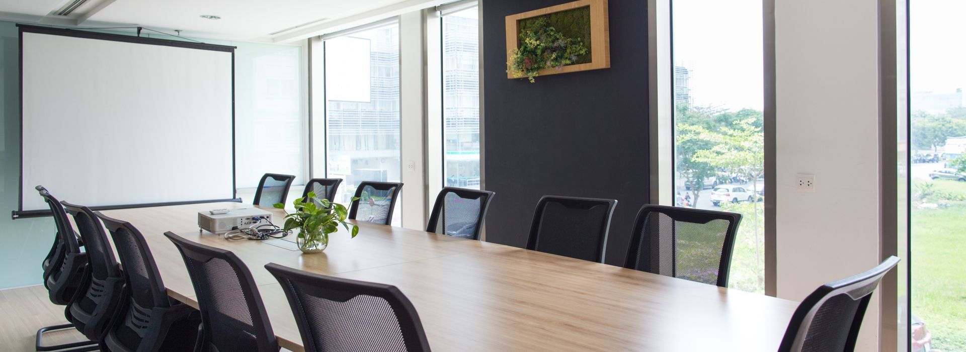 A modern conference room with a large table, black chairs, a projector screen, and a window overlooking a green space.