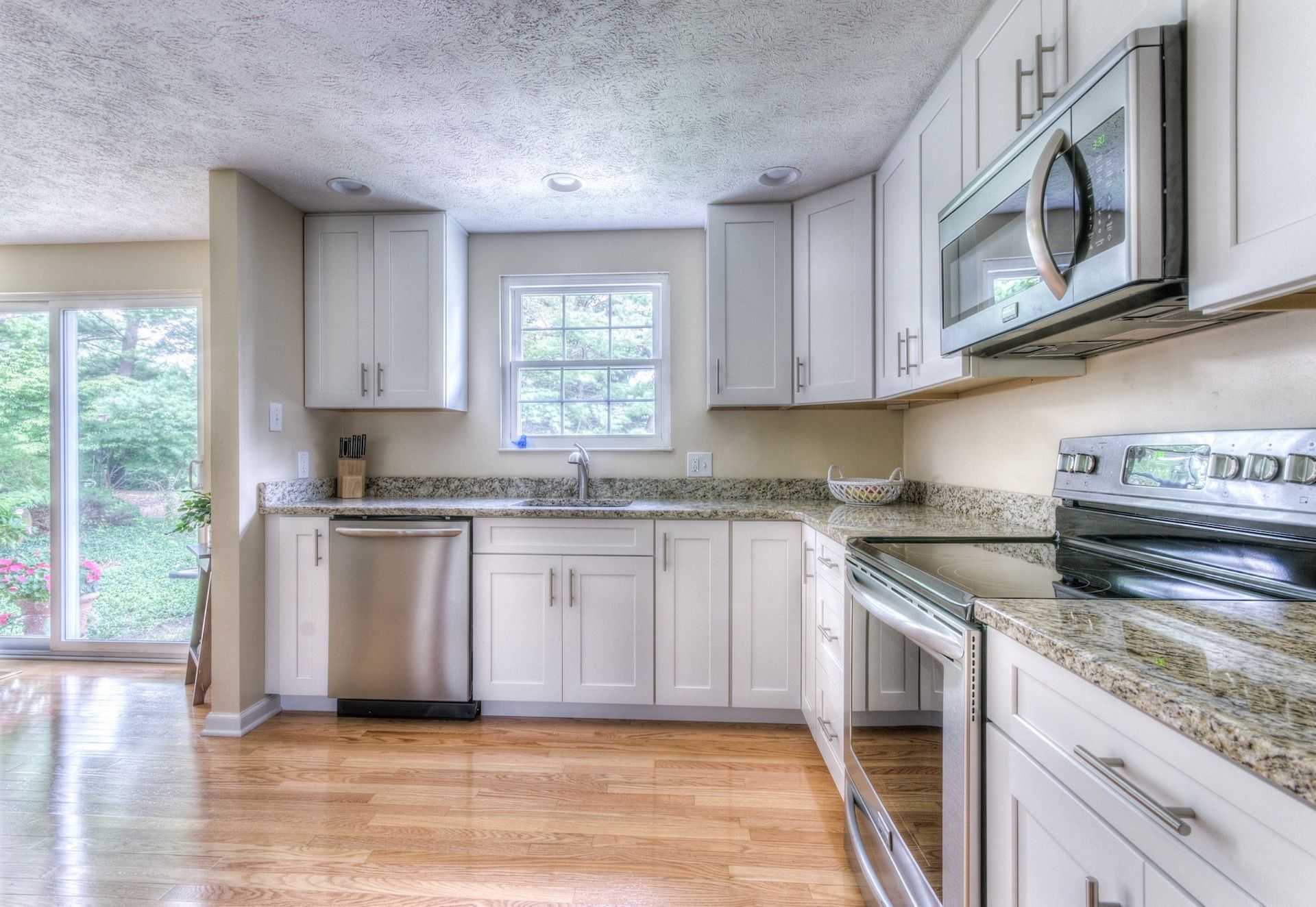 Bright, modern kitchen with white cabinets, stainless steel appliances, and granite countertops. Hardwood floors and a view of greenery through a sliding glass door.