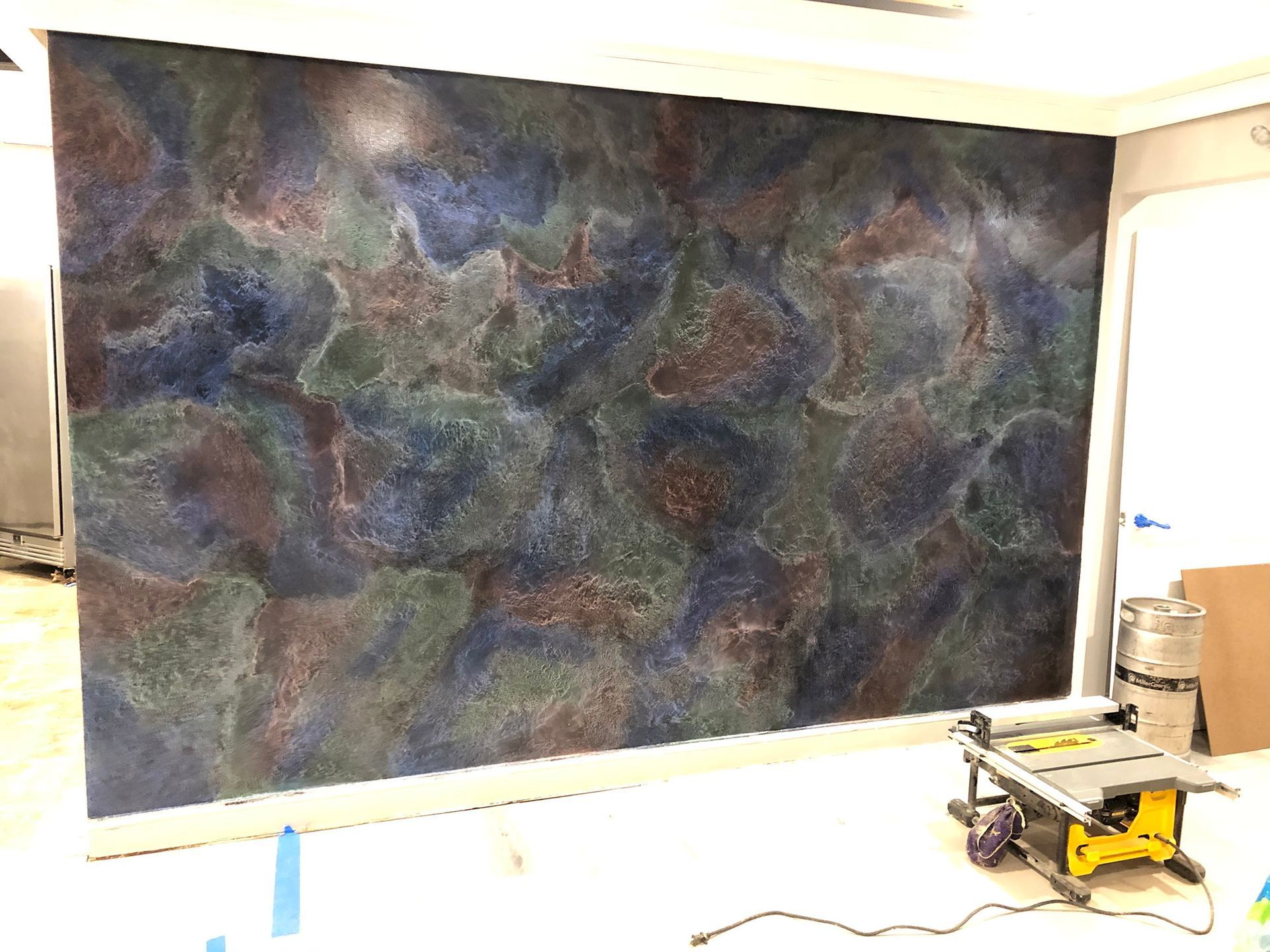 Large wall with a mottled, swirling design in shades of blue, green, and brown. A table saw and other construction materials are in the foreground.