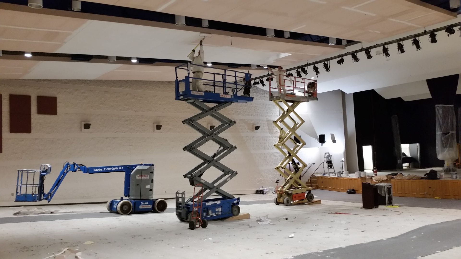 Construction site: three scissor lifts and a boom lift used for ceiling work. Workers are on the lifts. Interior space, walls are white.