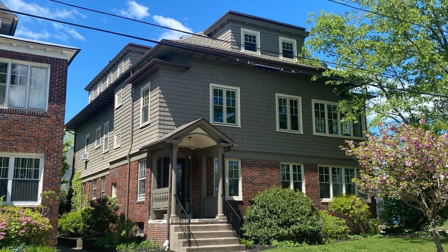 Three-story brick and gray building with a covered entrance, stairs, and small garden on a sunny day.
