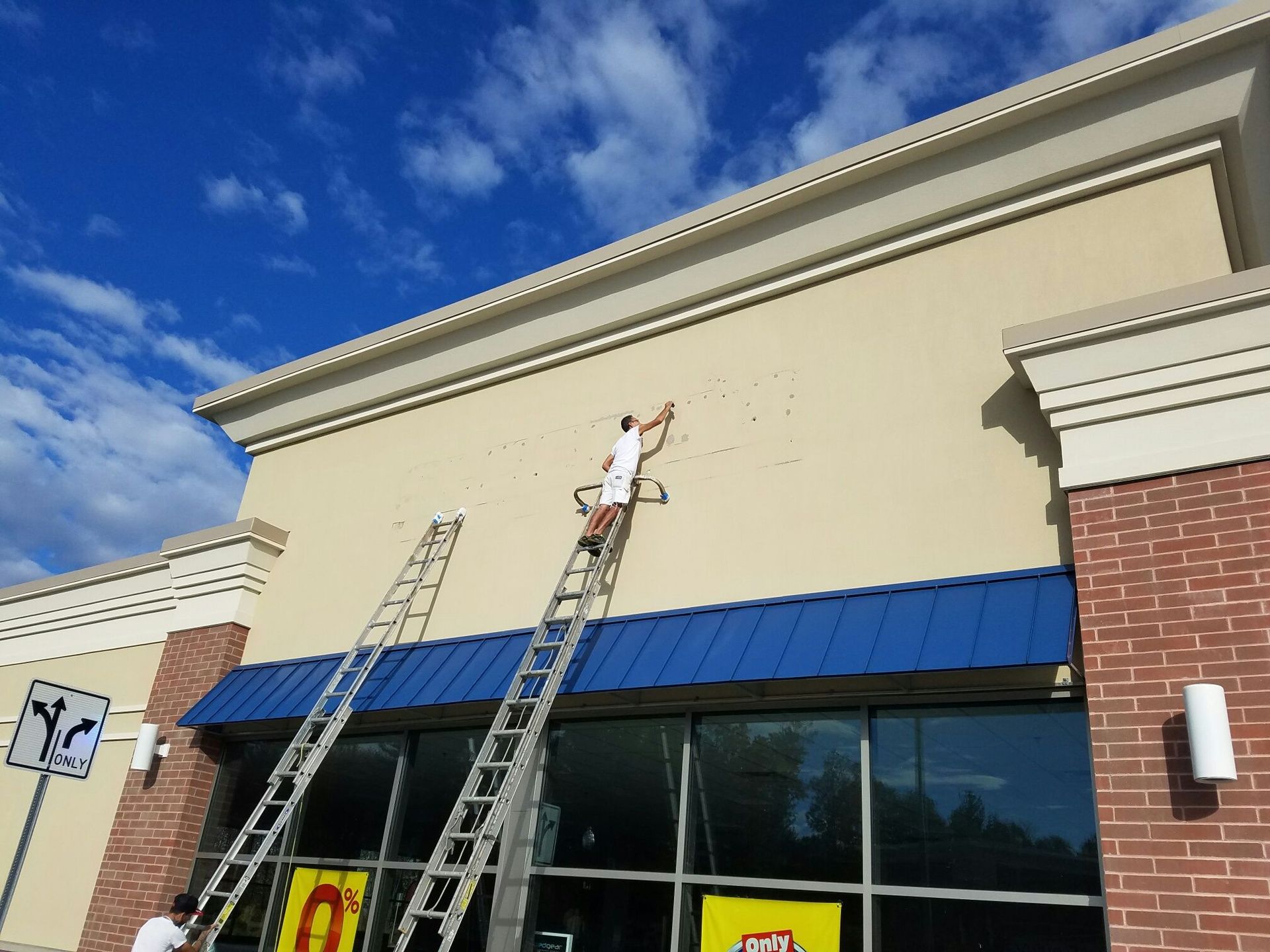 Two painters on ladders are working on the exterior of a commercial building. One is painting the tan facade.