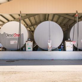 Large cylindrical fuel tanks under a shelter with visible fuel pumps. The tanks are white, one labeled