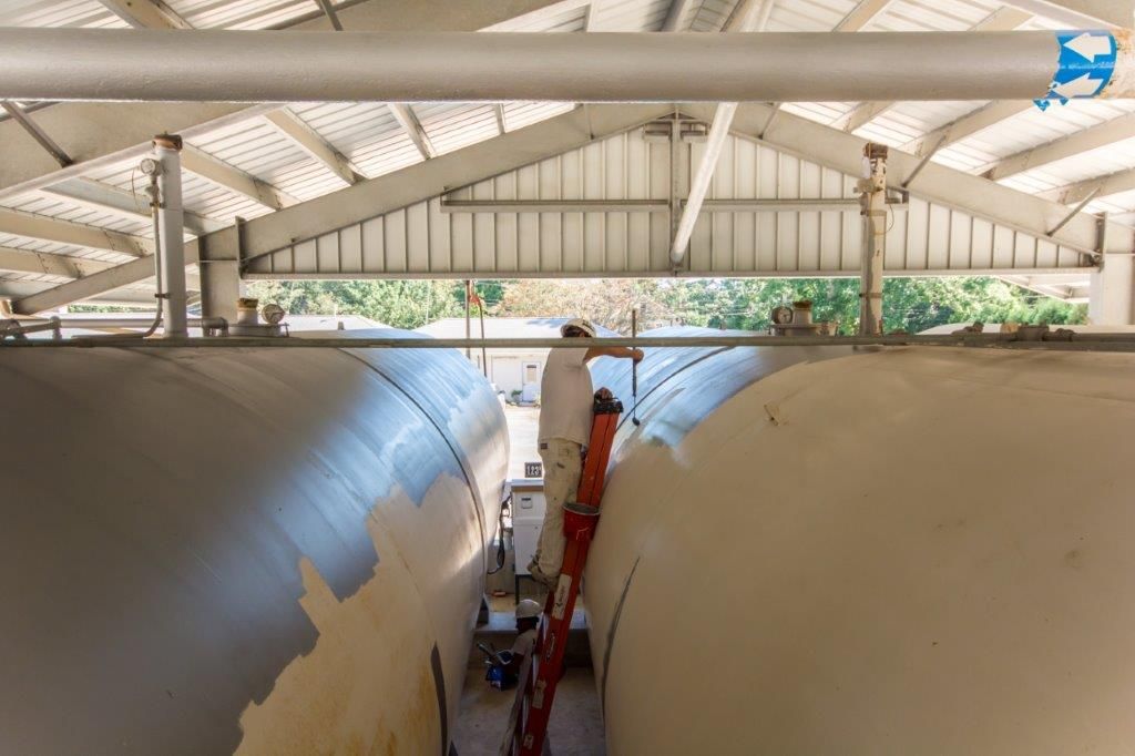 Person painting large metal tanks inside a warehouse. The person is standing on a ladder.