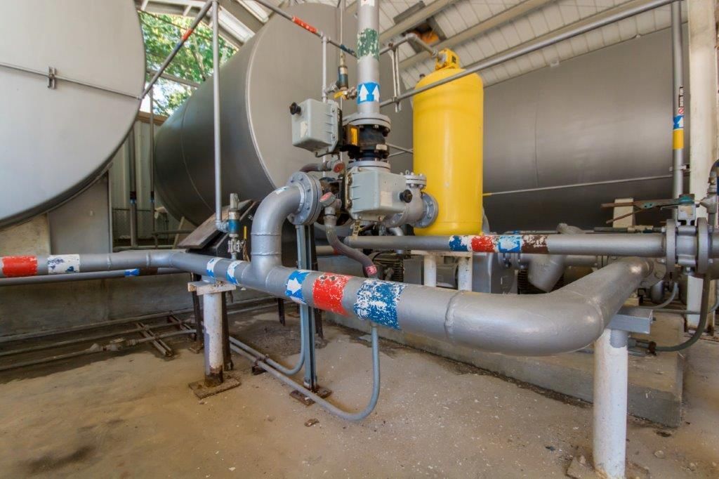 Industrial piping and tanks in a storage facility. Gray pipes with blue and red markings are in front of large silver tanks; a yellow cylinder is also visible.